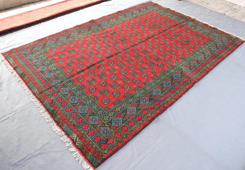 Op de afbeelding: Een rood tapijt met een geometrisch patroon in tinten van groen en blauw. Het tapijt heeft een franje rand.
