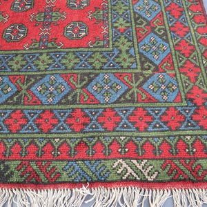 Op de afbeelding: Een rood, groen en blauw patroonteppich met een franje rand. Het tapijt heeft een geometrisch ontwerp met herhalende motieven.