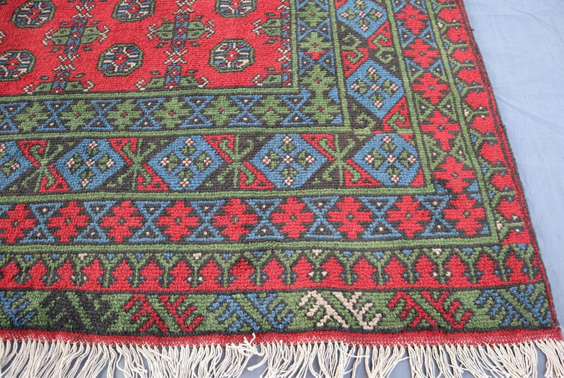 Op de afbeelding: Een rood, groen en blauw patroonteppich met een franje rand. Het tapijt heeft een geometrisch ontwerp met herhalende motieven.