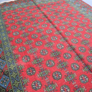 Op de afbeelding: Een rood tapijt met een geometrisch patroon in tinten blauw en groen. Het tapijt heeft een rand met een ingewikkelder ontwerp.