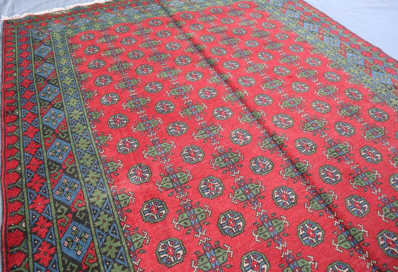 Op de afbeelding: Een rood tapijt met een geometrisch patroon in tinten blauw en groen. Het tapijt heeft een rand met een ingewikkelder ontwerp.