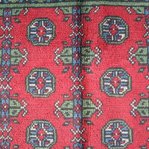 Op de afbeelding: Close-up van een rood tapijt met een herhalend patroon van groene, blauwe en witte geometrische vormen. Het tapijt heeft een traditioneel ontwerp.