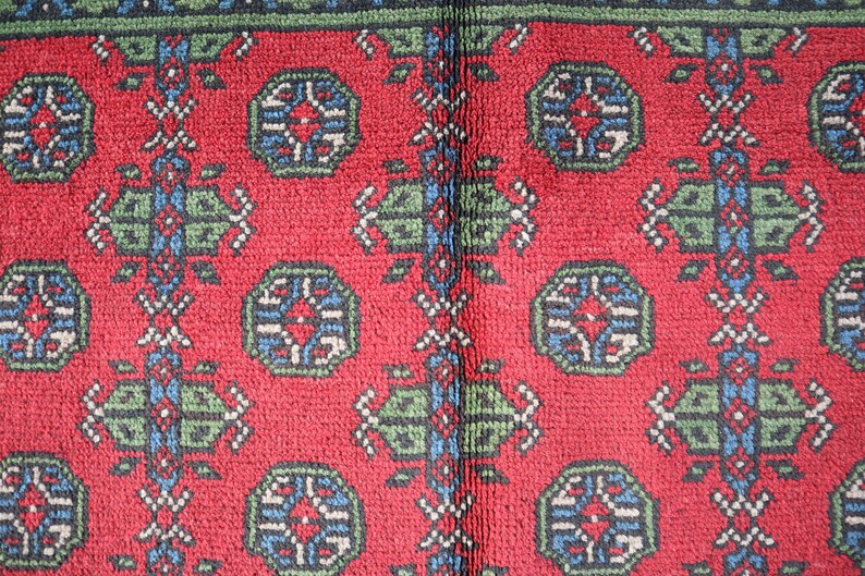 Op de afbeelding: Close-up van een rood tapijt met een herhalend patroon van groene, blauwe en witte geometrische vormen. Het tapijt heeft een traditioneel ontwerp.