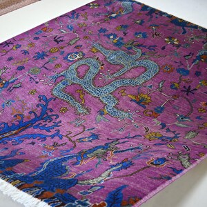 May include: A vibrant purple rug with a detailed design featuring a large, light blue dragon in the center. The rug has a complex pattern with blue, yellow, and green accents, and a white fringe along one edge. The rug is likely made of wool or a similar material.