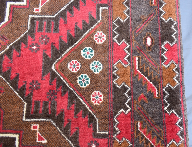 Puede incluir: Primer plano de una alfombra roja, marr&oacute;n y negra con un patr&oacute;n geom&eacute;trico. La alfombra tiene un borde blanco y presenta un dise&ntilde;o repetitivo de flores y diamantes.