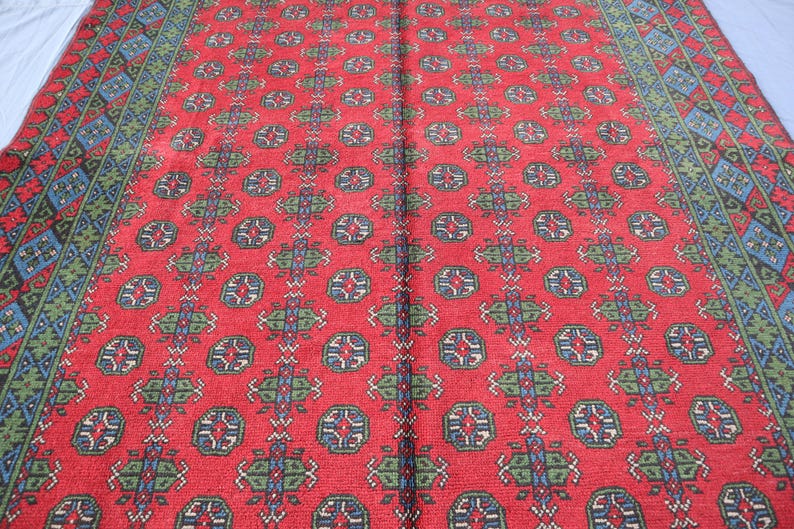 Op de afbeelding: Een rood tapijt met een herhalend patroon van blauwe, groene en witte geometrische vormen. Het tapijt heeft een rand met een vergelijkbaar patroon.