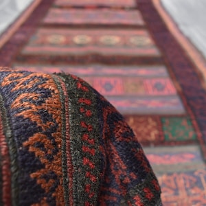 May include: Close-up of a colourful woven rug with intricate patterns in shades of red, brown, green, and blue. The rug has a fringed edge and is made of wool.