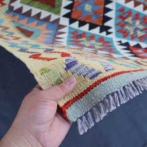 May include: A colorful woven rug with a geometric pattern in shades of red, blue, green, and yellow. The rug has a fringe border and is being held by a hand.