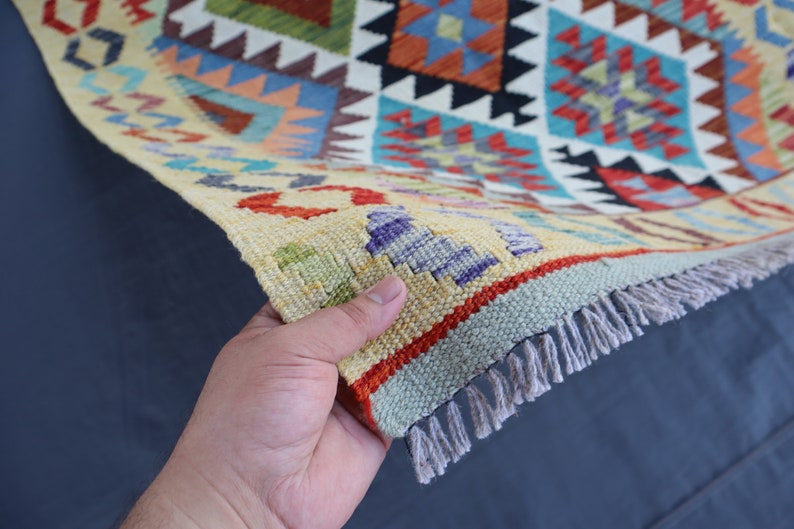 May include: A colorful woven rug with a geometric pattern in shades of red, blue, green, and yellow. The rug has a fringe border and is being held by a hand.