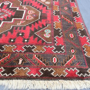 Puede incluir: Una alfombra roja, marr&oacute;n y azul con un patr&oacute;n y un borde de flecos. La alfombra tiene un dise&ntilde;o tradicional con formas geom&eacute;tricas y motivos florales.