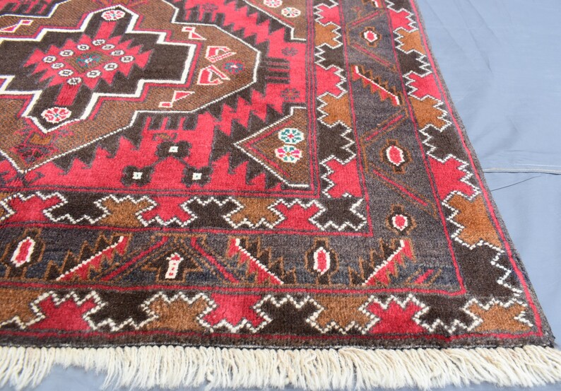 Puede incluir: Una alfombra roja, marr&oacute;n y azul con un patr&oacute;n y un borde de flecos. La alfombra tiene un dise&ntilde;o tradicional con formas geom&eacute;tricas y motivos florales.