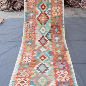 May include: A long, rectangular rug with a vibrant geometric pattern. The rug features a central column of diamond and rectangular shapes in red, orange, blue, and green hues, bordered by white and cream. The rug is likely made of woven fabric.