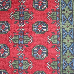 Op de afbeelding: Een close-up van een rood tapijt met een herhalend patroon van groene, blauwe en witte geometrische vormen. Het tapijt heeft een rand met een vergelijkbaar patroon.
