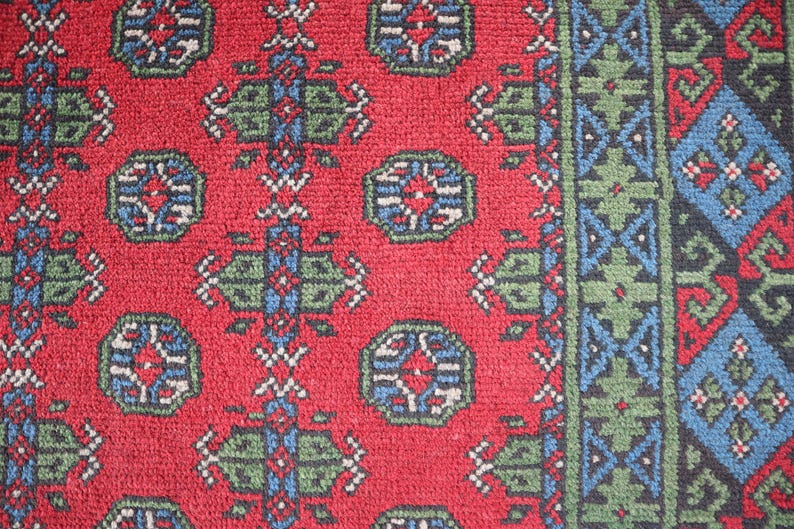 Op de afbeelding: Een close-up van een rood tapijt met een herhalend patroon van groene, blauwe en witte geometrische vormen. Het tapijt heeft een rand met een vergelijkbaar patroon.