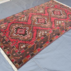 Puede incluir: Una alfombra roja, marr&oacute;n y blanca con un dise&ntilde;o geom&eacute;trico. La alfombra tiene un borde de flecos.