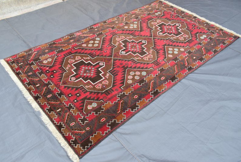 Puede incluir: Una alfombra roja, marr&oacute;n y blanca con un dise&ntilde;o geom&eacute;trico. La alfombra tiene un borde de flecos.