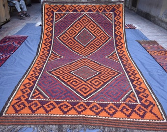 May include: A large, intricately patterned rug with a geometric design in shades of orange, brown, blue, and white. The rug is folded in half and laid out on a blue surface.