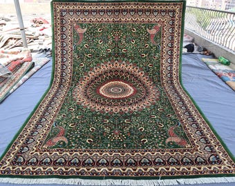Può includere: Un grande tappeto rettangolare verde con un intricato design floreale e paisley. Il tappeto presenta un medaglione centrale con un motivo dettagliato nei toni del rosso, marrone e crema. Il bordo include motivi di pavone e una frangia color crema.