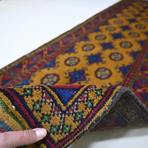 May include: A rectangular, hand-woven rug with a mustard yellow field and intricate geometric patterns in blue, red, and green. The rug's border features a repeating design, and the fringe is visible at one end. The rug is partially rolled up, revealing its underside.