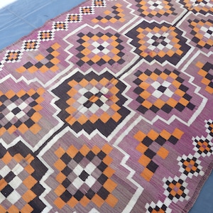 May include: A woven rug with a geometric pattern in shades of purple, orange, black, and white. The rug has a traditional design with repeating squares and diamonds.