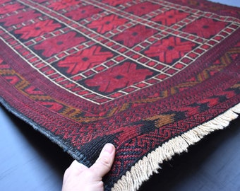 May include: A red, black, and white patterned rug with a fringe border. The rug is folded in half and held by a hand.
