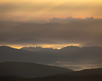 Könnte beinhalten: Ein Panoramablick auf eine Bergkette bei Sonnenaufgang, mit Schichten von Nebel und Wolken, die eine weiche, ätherische Atmosphäre erzeugen. Der Himmel ist in einem warmen, goldgelben Ton gehalten, und die Berge sind vor dem Licht silhouettiert.