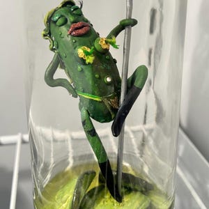 Könnte beinhalten: Eine skurrile Gurkenfigur in einem Glas, die auf einer silbernen Stange posiert. Die Gurke ist mit einer Perücke, Make-up und Accessoires verziert. Das Glas enthält grüne Flüssigkeit und steht auf einem weißen Regal.