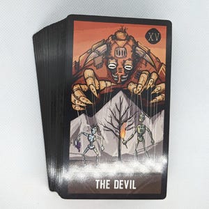 May include: A deck of tarot cards with black edges. The Devil card features a mechanical demon with puppet strings controlling two figures. The background is orange with a tree and the text "THE DEVIL".