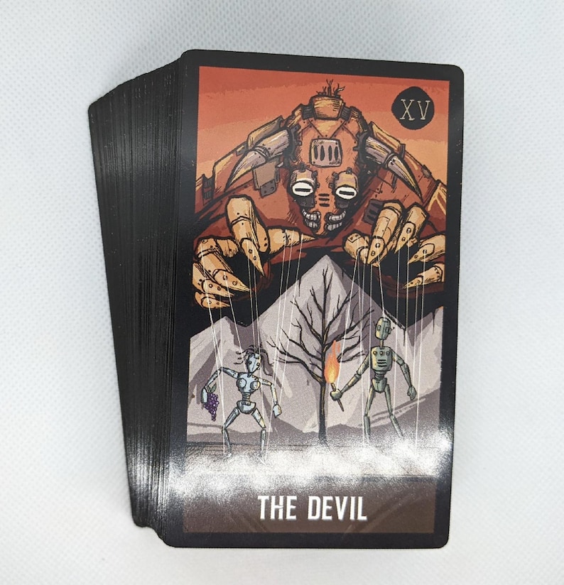May include: A deck of tarot cards with black edges. The Devil card features a mechanical demon with puppet strings controlling two figures. The background is orange with a tree and the text "THE DEVIL".