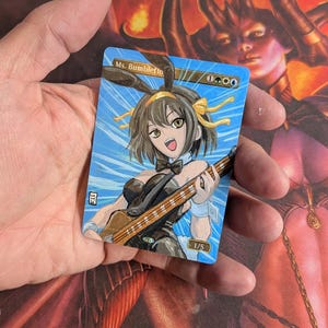 May include: A custom trading card featuring an anime-style character playing a bass guitar. The character, with bunny ears and a bow tie, is set against a blue sky background. The card is labeled "Ms. Bumbleflo" and includes mana symbols.