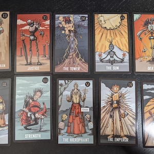 May include: A collection of ten tarot cards featuring stylized illustrations. Each card has a unique design and title, including "The Hanged Man," "Justice," "The Tower," and "The Sun." The cards have a dark border and a muted color palette.