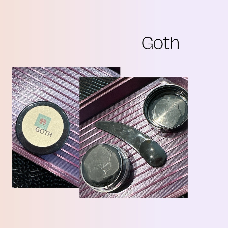 May include: A set of black beauty products in a purple and black striped container. The set includes a black jar with a black lid, a black jar with a silver lid, and a black facial tool. The jar with the silver lid has the word "GOTH" printed on it.