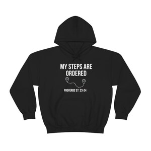 Op de afbeelding: Zwart hoodie met witte tekst die "Mijn stappen zijn geordend" en "Spreuken 37:23-24" luidt. De tekst wordt vergezeld door een afbeelding van een stippellijn met twee locatiepin-pictogrammen.