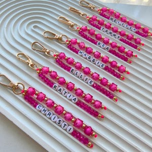 Könnte beinhalten: Eine Sammlung von sechs Schlüsselanhängern mit goldenen Karabinerverschlüssen, jeweils mit zwei Strängen aus leuchtend pinkfarbenen facettierten Perlen und kleinen goldenen Zwischenperlen. Ein Strang enthält weiße Buchstabenperlen, die die Namen MELISSA, TAMMY, BEA, MAGGIE, ELLIE, KATIA und CHRISTO bilden.