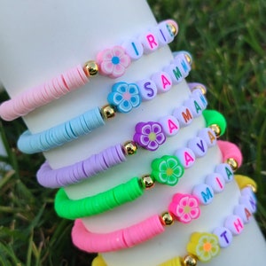 May include: A stack of colorful beaded bracelets. Each bracelet features a different color of flat beads, flower-shaped beads, and letter beads spelling out names. The bracelets are pink, blue, purple, green, pink, and yellow.