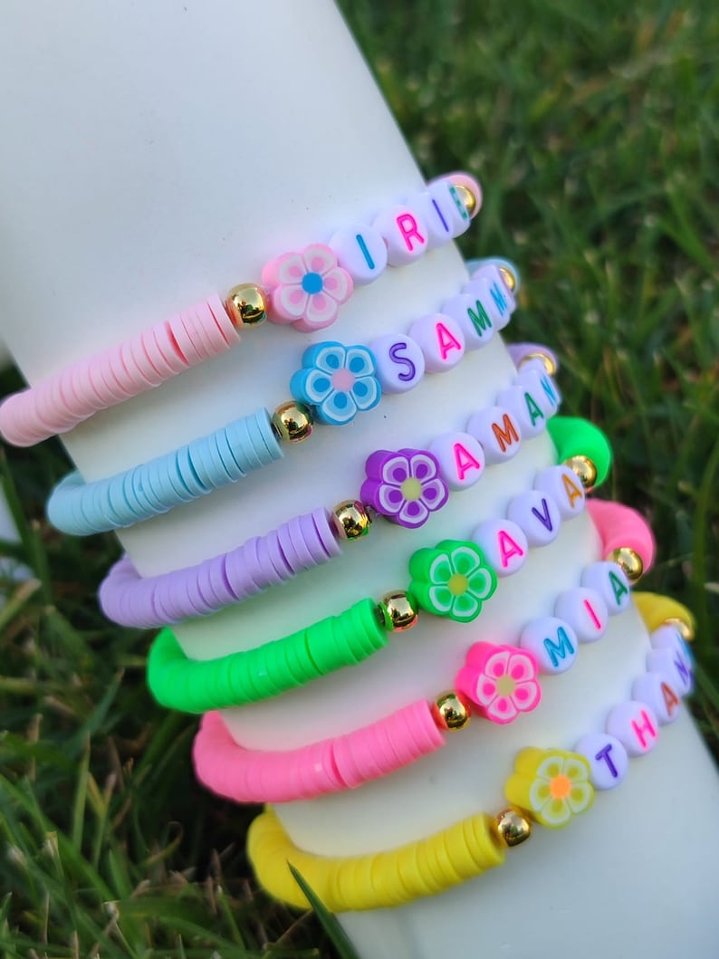 May include: A stack of colorful beaded bracelets. Each bracelet features a different color of flat beads, flower-shaped beads, and letter beads spelling out names. The bracelets are pink, blue, purple, green, pink, and yellow.