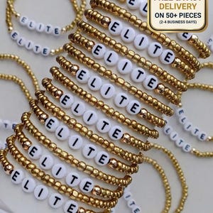 Puede incluir: Una colección de pulseras elásticas con pequeñas cuentas metálicas doradas y cuentas blancas circulares con el texto negro impreso ELITE. Las pulseras están dispuestas en una pila, mostrando el patrón repetitivo de cuentas doradas y la palabra ELITE.