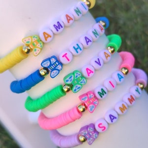 May include: A stack of colorful beaded bracelets with names spelled out in letter beads. Each bracelet features a different color of beads, including yellow, blue, green, pink, and purple. Butterfly charms and gold accents add detail to the bracelets.