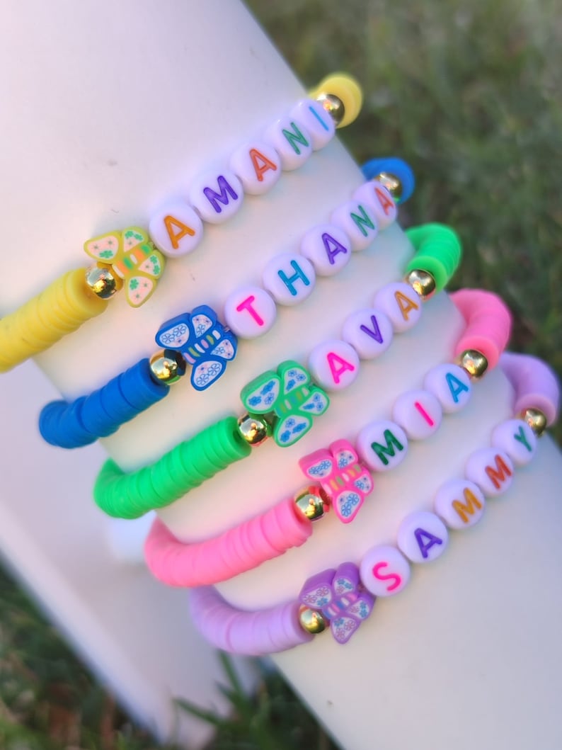 May include: A stack of colorful beaded bracelets with names spelled out in letter beads. Each bracelet features a different color of beads, including yellow, blue, green, pink, and purple. Butterfly charms and gold accents add detail to the bracelets.