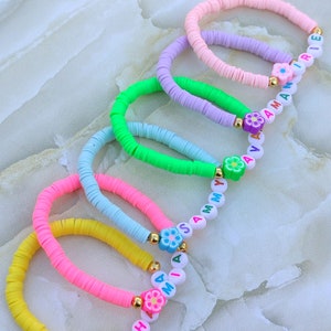 May include: A collection of colorful beaded bracelets. The bracelets feature flat, disc-shaped beads in shades of yellow, pink, blue, green, and purple. Each bracelet has a flower charm and some have letter beads spelling out names. Gold-colored beads are used as accents.