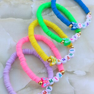 May include: A collection of colorful beaded bracelets. Each bracelet features a different color, including blue, green, yellow, pink, and purple. The bracelets have gold accents and include names spelled out with white letter beads. The bracelets are a fun accessory.