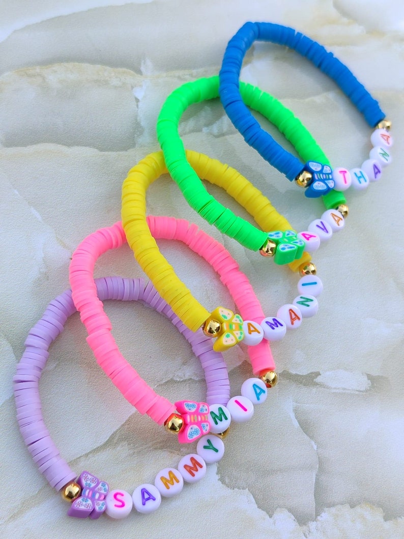 May include: A collection of colorful beaded bracelets. Each bracelet features a different color, including blue, green, yellow, pink, and purple. The bracelets have gold accents and include names spelled out with white letter beads. The bracelets are a fun accessory.