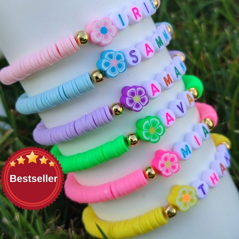 May include: A stack of colorful beaded bracelets. Each bracelet features a different color of flat beads, gold accents, and flower-shaped beads. The bracelets spell out names like "IRIE", "SAMAN", "AVA", "MIA", and "THAN".