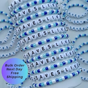 May include: A stack of elastic bracelets made with white, light blue, and dark blue beads. Each bracelet features white letter beads spelling out the phrase C SAVES LIVES, interspersed with small silver-toned metallic spacer beads throughout.