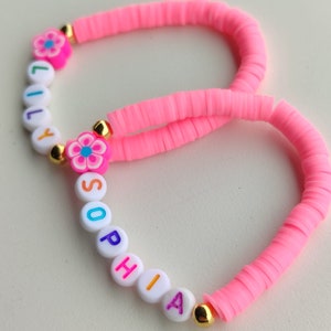 May include: Two handmade bracelets. One is inscribed with the name "LILY" and the other with "SOPHIA". Both bracelets are made of bright pink, flat, disc-shaped beads. Each bracelet features a flower charm and gold-colored accents.