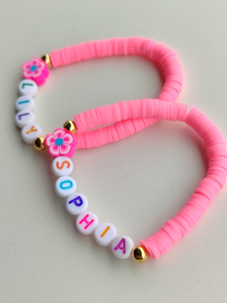 May include: Two handmade bracelets. One is inscribed with the name "LILY" and the other with "SOPHIA". Both bracelets are made of bright pink, flat, disc-shaped beads. Each bracelet features a flower charm and gold-colored accents.