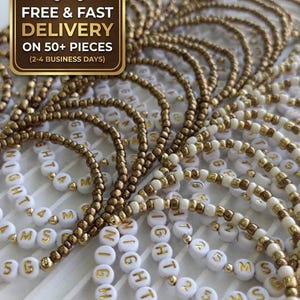 May include: A collection of beaded bracelets featuring metallic gold-toned beads and white circular beads with gold lettering. The white beads display the text NIGHT 25 MS, arranged in repeating patterns across multiple strands of jewellery.