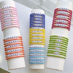 Puede incluir: Tres expositores cilíndricos con pilas de pulseras de cuentas. La pila izquierda presenta cuentas rosas y naranjas con el texto JIN y J-HOPE. La pila central tiene cuentas azules, amarillas y azul claro con el texto RM, JIMIN y JUNGKOOK. La pila derecha tiene cuentas rojas y verdes con el texto SUGA y V.