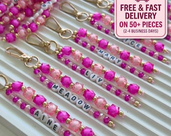 May include: A collection of beaded keychains featuring gold-toned lobster clasps and strands of vibrant pink, light pink, and clear beads. Each keychain includes white letter beads spelling out names like Meadow, Liv, Mary, and Aine.
