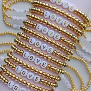 May include: A stack of gold beaded bracelets with white beads that spell out "LOVE".